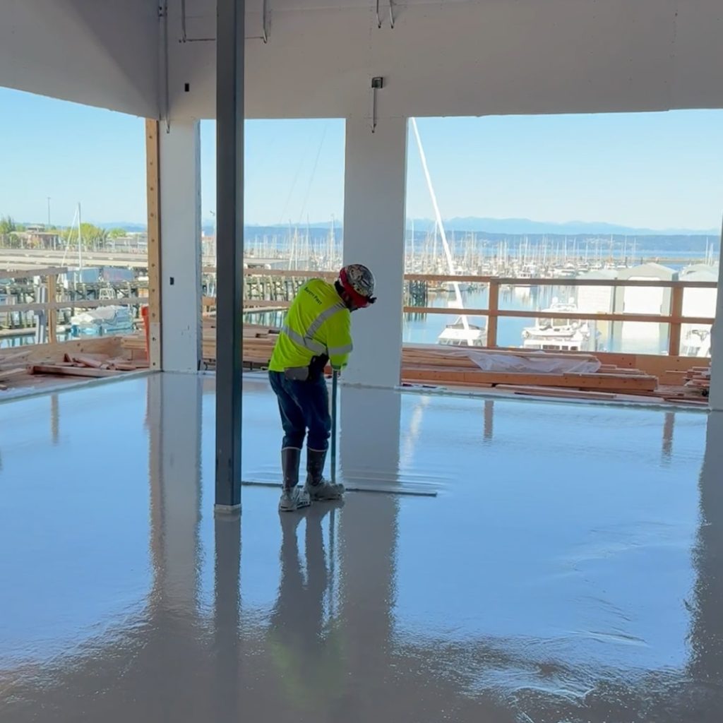 Worker in yellow vest smoothing glossy concrete floor with long squeegee at waterfront building